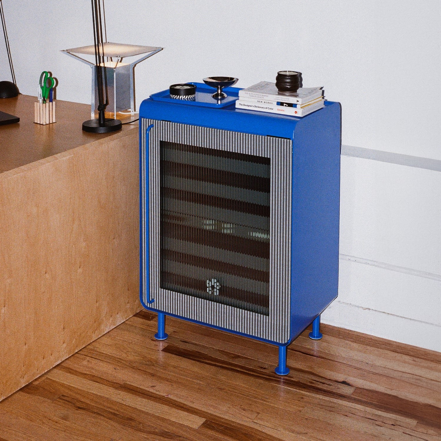 Blue Rocco Classic drinks fridge with a reeded glass door and a tray on top, beside a plywood desk